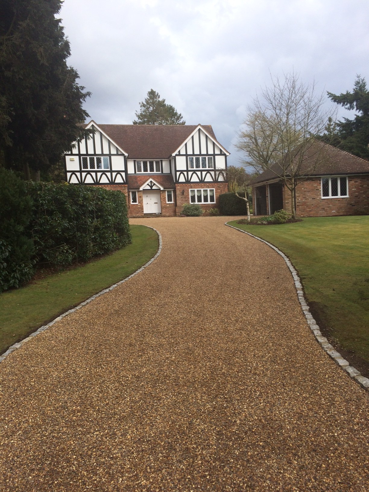 A long curving golden gravel drive with stone edging leading to a Tudor style house